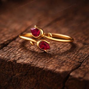 May include: A gold-colored ring featuring a floral design with two teardrop-shaped, deep red gemstones. The ring is set against a textured, dark brown wooden surface, creating a contrast between the shiny metal and the natural background.