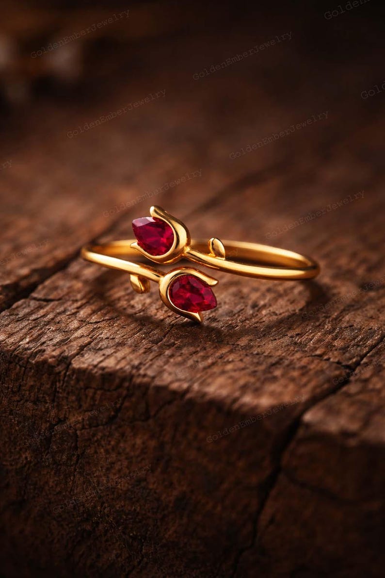May include: A gold-colored ring featuring a floral design with two teardrop-shaped, deep red gemstones. The ring is set against a textured, dark brown wooden surface, creating a contrast between the shiny metal and the natural background.
