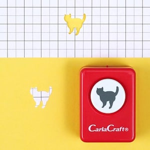 Op de afbeelding: Een rode CarlaCraft® katvormige papierpons met een grijze kattenomtrek op een witte cirkel. Een gele katvormige uitsparing en een katvormige papieren uitsparing zijn ook zichtbaar tegen een gele en rasterachtergrond.