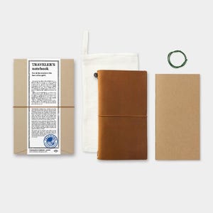May include: A Traveler's Notebook set includes a brown leather journal, a kraft paper refill, a canvas pouch, and a green elastic band. The journal is packaged in a brown cardboard sleeve with text and a label that reads "Traveler's Notebook".