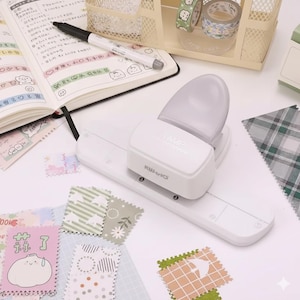 May include: A white stamp maker with the text "STAMP" and "KW-trio" is surrounded by colourful patterned paper squares, a notebook, a pen, and decorative tape rolls. The stamp maker is on a white surface.