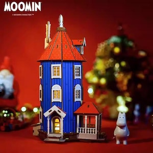 May include: A wooden model of a blue house with a red roof, white window frames, and a small porch. The house has a tall, cylindrical shape with multiple windows. A white figure stands to the right of the house. The word "MOOMIN" is at the top left.