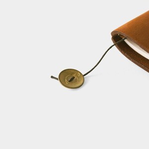 May include: A close-up of a brown leather journal with a brass button closure. The button is round with a small design and is attached to the journal with a dark green string. The journal's pages are visible.