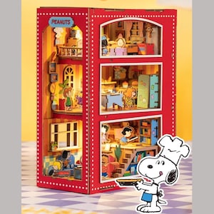 Op de afbeelding: Een rood, drieledig poppenhuis met witte stippen en het woord "PEANUTS" bovenaan. Het poppenhuis heeft miniatuurkamers met Snoopy en andere Peanuts-personages. Snoopy, gekleed als chef-kok, houdt een taart vast.