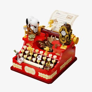 May include: A red toy typewriter featuring Snoopy and Woodstock characters. The typewriter has a clock, a sheet of paper, and a small gramophone. The word "Typewriter" is printed on the front. The keys are cream-coloured with letters and paw prints.