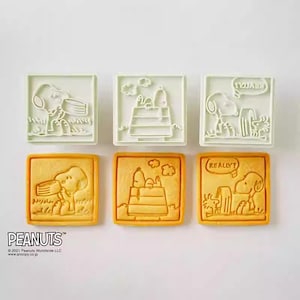 six square cookie cutters shaped like characters from the Peanuts comic strip, including Snoopy, Woodstock, and other iconic figures.