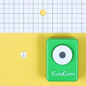 May include: A green paper punch with a white and black center, labeled "CarlaCraft®", sits on a yellow surface. A yellow circle and a circle with a cross are also visible. The background is a white grid.