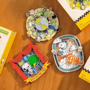 May include: Three colorful Snoopy-themed magnets. One is a circular design with Snoopy and friends around a globe. Another features Snoopy in a biplane with the word "Fly". The third shows Snoopy with a kite-eating tree. The magnets are on a wooden surface.