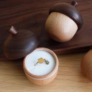 May include: A gold ring with a cat charm and a small coin charm sits in a wooden acorn-shaped ring box. The box is made of light wood with a dark brown top. Two other acorn boxes are visible.