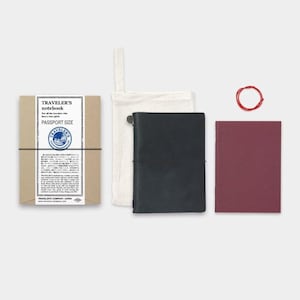 May include: A Traveler's Notebook passport size set. The set includes a brown paper box with text, a black leather notebook, a burgundy notebook insert, a white canvas bag, and a red elastic band. The items are arranged on a white background.