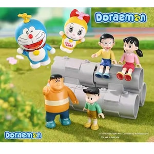 May include: A collection of colourful Doraemon character figurines. The figures include Doraemon, a yellow character with a red bow, and other characters. The figures are posed on a grey cylindrical structure, with the Doraemon logo in the background.