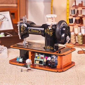 Puede incluir: Modelo de máquina de coser negra y dorada con la palabra "Pantasy" en el lateral. El modelo tiene una base de madera con una pequeña figura de una persona delante. Se ven carretes de hilo en el fondo.