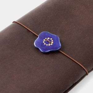 Könnte beinhalten: Ein dunkelbraunes Lederjournal mit einem blauen Blumenknopfverschluss. Der Knopf hat eine goldene Mitte und goldene Punkte am Rand. Eine braune elastische Kordel sichert das Journal.