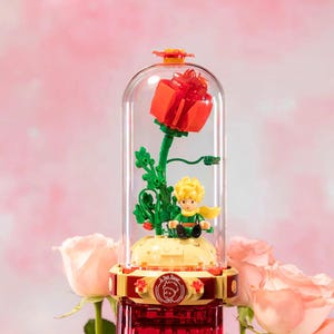 May include: A glass dome ornament with a red rose and a small figure of a boy with blond hair and a yellow scarf. The base is yellow and red, with a circular emblem. The background is a soft pink.