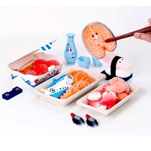 Op de afbeelding: Een verzameling pluche sushi-speelgoed, waaronder zalm, garnalen en nigiri, gerangschikt in serveerbakjes en een afhaalbox. Een blauwe sake-fles en een klein bekertje zijn ook aanwezig. Eetstokjes houden een stuk zalm vast.