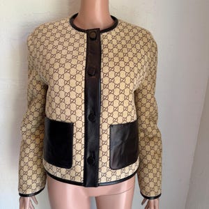 May include: A beige jacket featuring a brown repeating GG logo pattern, accented with dark brown leather trim along the collar, cuffs, and hem. The jacket includes a central vertical leather placket with four matching buttons and two large leather pockets.