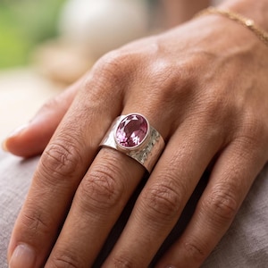 May include: A wide silver band ring featuring a central oval-cut pink gemstone. The metal surface of the ring has a hammered, textured finish, and the stone is secured in a smooth bezel setting with a delicate beaded edge detail around the perimeter.