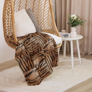 May include: A soft throw blanket featuring a realistic tiger stripe pattern in shades of orange, black, and white. The blanket is draped over a woven rattan hanging chair, which also contains a white textured pillow and a grey pillow.