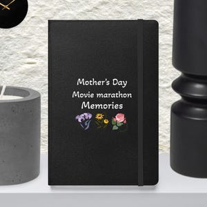 May include: A black journal with the text "Mother's Day Movie marathon Memories" and floral illustrations. The journal has an elastic band closure. The background is a neutral color.