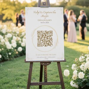 May include: A wooden easel displays a sign with the text "Help Us Capture the Magic" and a QR code. The sign also includes the names "EMMA & JAMES" and the date "06-26-2026". The sign is set up outdoors on a grassy area.