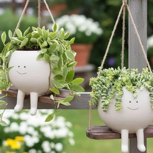 Może przedstawiać: Dwie wiszące donice o fantazyjnym designie. Każdy biały ceramiczny garnek ma uśmiechniętą twarz i nogi, zawieszony na sznurku. Zwieszające się zielone rośliny wylewają się z krawędzi, tworząc wesoły, dekoracyjny akcent.