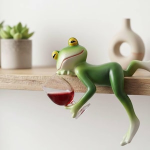 May include: A whimsical green frog figurine holding a wine glass filled with red wine. The frog is resting on a wooden shelf. The frog has yellow eyes and white accents. The wine glass is clear. Decorative item.