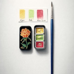Può includere: Un set di acquerelli con un pennello con manico blu. Il set include una scatola nera con un disegno di rosa, una scatola con tre acquerelli e una tavolozza di colori con le parole "light", "like" e "Rose blush".