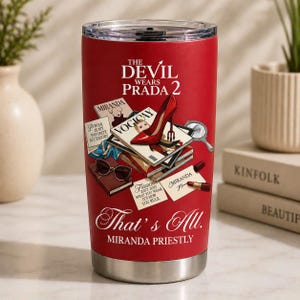 May include: A red stainless steel tumbler featuring illustrations and text from "The Devil Wears Prada 2." The design includes a red high heel, fashion magazines, and the quote "That's All. Miranda Priestly."