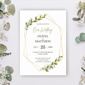 Op de afbeelding: Een trouwkaart met een gouden geometrisch kader en groene bladaccenten. De uitnodiging luidt "Our Wedding" met de namen Olivia & Matthew en de datum 25 juli 2026. De uitnodiging is omgeven door groen en bloemen.