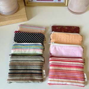 May include: A collection of eight small rectangular fabric pouches arranged in two columns. The pouches feature various patterns including horizontal stripes, polka dots, and floral prints in colours such as pink, yellow, brown, black, and white.