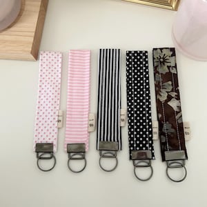 May include: Five fabric wristlet keychains with metal hardware and circular rings. The patterns include pink polka dots, pink stripes, black and white vertical stripes, black with white polka dots, and a dark brown floral print. Each has a small tag labelled IS.