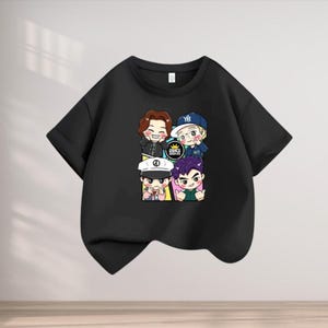 Puede incluir: Camiseta negra de manga corta con un gráfico de dibujos animados de cuatro personajes sonrientes. El gráfico incluye el texto "BIG BANG" en un emblema dorado. Los personajes tienen diferentes colores de pelo y usan varios accesorios.