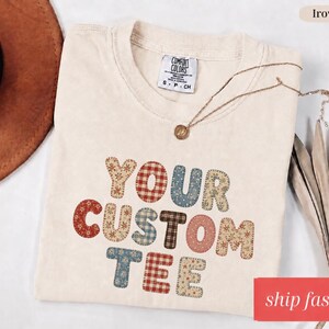 May include: Ivory-colored Comfort Colors t-shirt with the words "YOUR CUSTOM TEE" in a patchwork design. The shirt has a crew neck and short sleeves. A gold necklace with a pendant is draped across the shirt. The words "ship fast" are in a red rectangle.