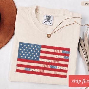 Op de afbeelding: Crèmekleurig T-shirt met een opgenaaid Amerikaans vlagontwerp. De vlag heeft blauwe sterren en rood-witte strepen. Het shirt heeft een ronde hals en een klein label met de tekst "Comfort Colors". Een gouden ketting is over het shirt gedrapeerd.