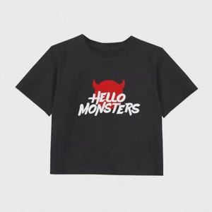 Puede incluir: Top corto negro con un gráfico de diablo rojo y el texto "Hello Monsters" en blanco. La camiseta tiene mangas cortas y cuello redondo. El diseño presenta una cabeza de diablo rojo con cuernos sobre el texto.