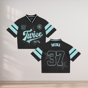 May include: Black cropped jersey with light blue accents. The jersey features the word "Twice" in a script font, the number "37", and the text "This Is For". The back of the jersey has the name "Mina" and the number "37".