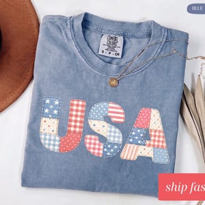 Op de afbeelding: Een blauw spijkerbroekkleurig t-shirt met het woord "USA" op de voorkant in een patchwork-ontwerp van rode, witte en blauwe stoffen. Een gouden ketting rust op het shirt. Het shirt is gelabeld "Comfort Colors".