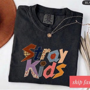 Op de afbeelding: Zwart T-shirt met de tekst "Stray Kids" in een patchwork ontwerp. Het shirt heeft een ronde hals en korte mouwen. Een gouden ketting met een hanger is over het shirt gedrapeerd. De woorden "ship fast" staan in een rode doos.