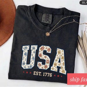 Op de afbeelding: Zwart T-shirt met het woord "USA" in een bloemenpatroon en de tekst "EST. 1776". Het shirt is van het merk Comfort Colors. Een gouden ketting ligt over het shirt.