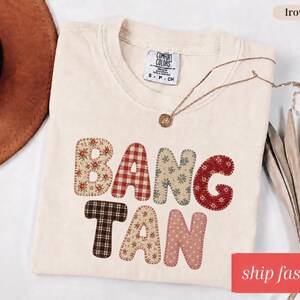 Op de afbeelding: Ivoor Comfort Colors T-shirt met het woord "BANGTAN" in patchwork letters. De letters hebben bloemen-, ruit- en stippatronen. Een gouden ketting met een hanger is over het shirt gedrapeerd. De woorden "ship fast" staan in een rode doos.