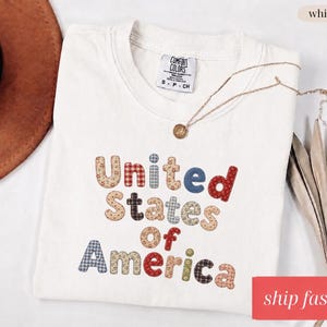 Op de afbeelding: Crèmekleurig T-shirt met de tekst "United States of America" in kleurrijke letters in patchwork-stijl. Een gouden ketting ligt op het gevouwen shirt. Een bruine hoed en gedroogde planten zijn op de achtergrond.