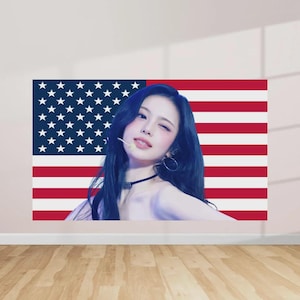 May include: A large wall art piece featuring the American flag as a backdrop. A person with dark hair and a black choker is positioned in front of the flag, winking at the viewer. The artwork is mounted on a white wall above a wooden floor.