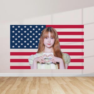 May include: A young person with long, light brown hair and a light green top forms a heart shape with their hands in front of an American flag. The flag is red, white, and blue, with stars in the upper left corner and stripes.