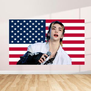 Könnte beinhalten: Ein Wandkunstdruck mit einer Person vor der amerikanischen Flagge. Die Person trägt ein weißes Hemd und eine schwarze Jacke mit Text. Der Hintergrund ist eine weiße Wand und ein Holzboden.