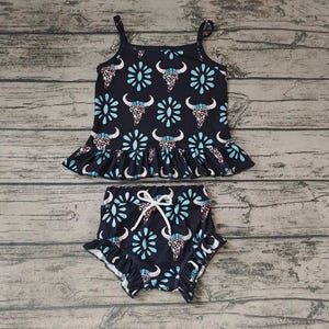 May include: A two-piece navy blue outfit featuring a tank top and high-waisted shorts. The set is adorned with a repeating pattern of bull skulls, turquoise floral accents, and a ruffled hem. The shorts have a white drawstring.