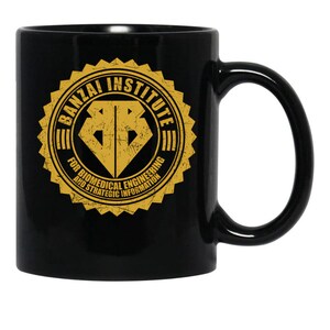 May include: Black ceramic mug featuring a gold emblem with the text "BANZAI INSTITUTE" and "FOR BIOMEDICAL ENGINEERING AND STRATEGIC INFORMATION." The emblem has a diamond-shaped design in the center.