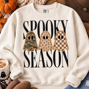Puede incluir: Una sudadera de cuello redondo color crema con el texto negro SPOOKY SEASON y tres ilustraciones de fantasmas. Los fantasmas tienen patrones distintos: uno con camuflaje verde, uno con estampado de leopardo beige y otro con cuadros marrones.