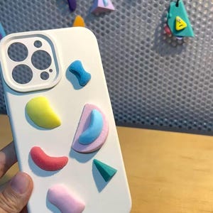 May include: White phone case with colorful, textured, abstract shapes in yellow, red, pink, blue, and green. The case has a camera cutout and is held by a hand. Additional colorful shapes are attached to a metal grid in the background.