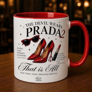 May include: White ceramic mug with a red interior and handle. The mug features the text "THE DEVIL WEARS PRADA 2" along with illustrations of red high heels, sunglasses, and lipstick. Additional text includes quotes and the phrase "That is All."