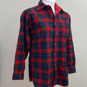 May include: A red, navy blue, and green plaid flannel shirt with a button-down front and a single chest pocket. The shirt has long sleeves and a classic collar. The inside collar is red. The shirt is displayed on a mannequin.